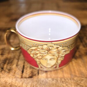 Puede incluir: Una taza de t&eacute; de porcelana con un exterior rojo intenso y un emblema central de una cabeza de Medusa dorada. La taza incluye un asa de color dorado, un interior blanco y un patr&oacute;n de greca en blanco y oro a lo largo del borde superior.