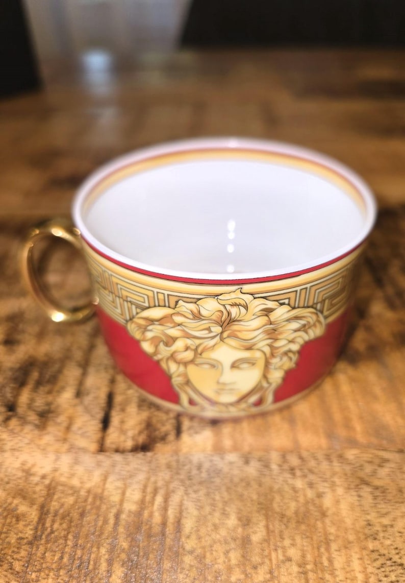 Puede incluir: Una taza de t&eacute; de porcelana con un exterior rojo intenso y un emblema central de una cabeza de Medusa dorada. La taza incluye un asa de color dorado, un interior blanco y un patr&oacute;n de greca en blanco y oro a lo largo del borde superior.