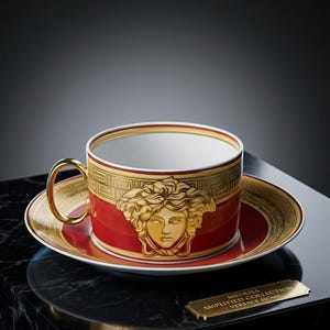 Puede incluir: Un juego de taza de t&eacute; y platillo de porcelana roja y dorada con un prominente emblema de cabeza de Medusa en oro. El borde est&aacute; decorado con un patr&oacute;n de greca dorada. Una peque&ntilde;a placa dorada dice MEDUSA AMPLIFIED COLLECTION - VERSACE HOME.