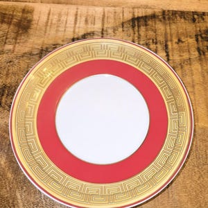 Puede incluir: Un plato redondo de cer&aacute;mica con un centro blanco, una banda roja ancha y un borde exterior decorado con un patr&oacute;n de greca en tono dorado. El plato tiene un borde fino de color oro y un acabado liso y brillante con motivos geom&eacute;tricos.