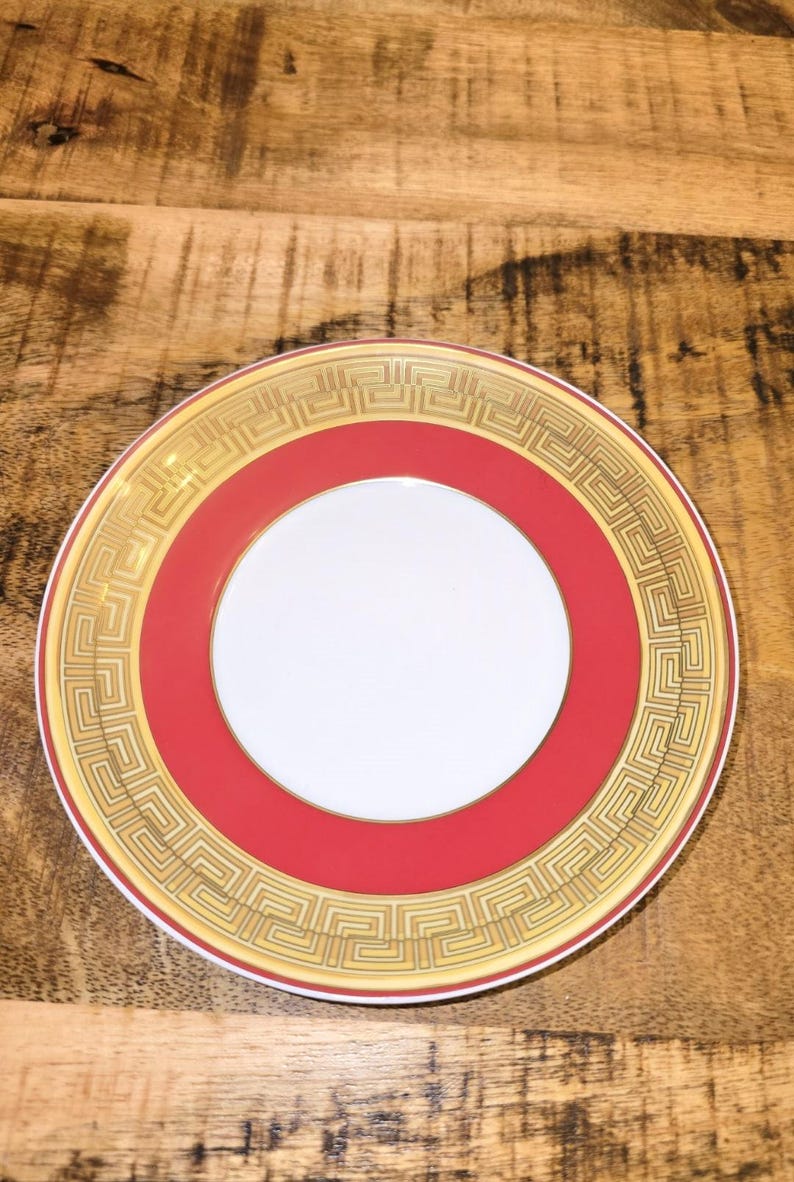 Puede incluir: Un plato redondo de cer&aacute;mica con un centro blanco, una banda roja ancha y un borde exterior decorado con un patr&oacute;n de greca en tono dorado. El plato tiene un borde fino de color oro y un acabado liso y brillante con motivos geom&eacute;tricos.