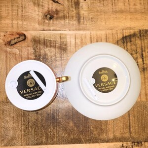 Puede incluir: Un juego de taza y platillo de porcelana blanca con asa dorada y etiquetas circulares negras en la parte inferior. Las etiquetas muestran el texto Versace, Medusa Amplified Golden Coin y el logotipo de Rosenthal. Vistos desde abajo.
