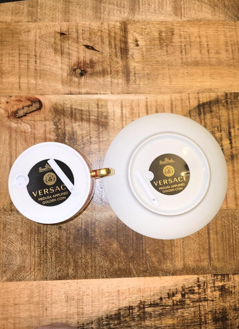 Puede incluir: Un juego de taza y platillo de porcelana blanca con asa dorada y etiquetas circulares negras en la parte inferior. Las etiquetas muestran el texto Versace, Medusa Amplified Golden Coin y el logotipo de Rosenthal. Vistos desde abajo.