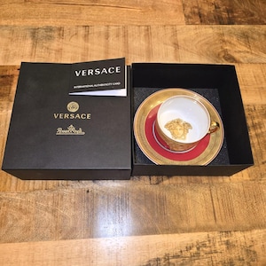 Puede incluir: Un juego de taza y plato de porcelana Versace y Rosenthal con un patr&oacute;n de greca en rojo y dorado y una cabeza de Medusa dorada en el interior de la taza. El conjunto se presenta en una caja negra con una tarjeta de autenticidad Versace.