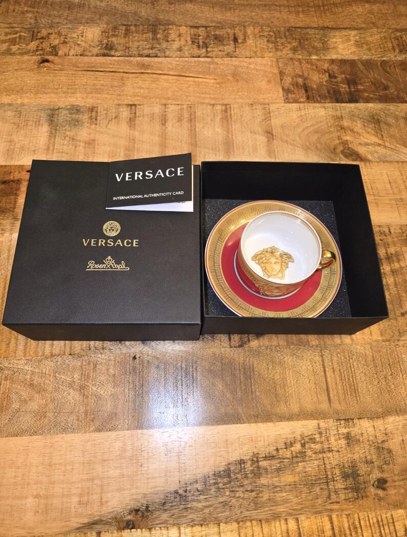 Puede incluir: Un juego de taza y plato de porcelana Versace y Rosenthal con un patr&oacute;n de greca en rojo y dorado y una cabeza de Medusa dorada en el interior de la taza. El conjunto se presenta en una caja negra con una tarjeta de autenticidad Versace.