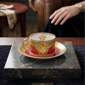 Puede incluir: Un juego de taza de t&eacute; y platillo de porcelana en rojo y oro, con un emblema de cabeza de Medusa dorada en el exterior e interior. La taza cuenta con un asa dorada y un borde con patr&oacute;n de greca. El conjunto tiene un acabado esmaltado.