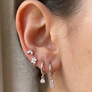 Könnte beinhalten: Nahaufnahme eines Ohrs, das mit mehreren silbernen Ohrringen geschmückt ist. Die Ohrringe umfassen kleine Ohrstecker mit Blatt- und Blumenmotiven sowie zwei Creolen, eine mit einem hängenden runden Edelstein und die andere mit einem rechteckigen Edelstein.