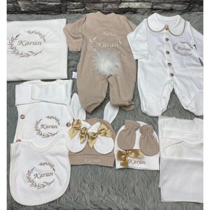May include: A baby clothing set in cream and beige. The set includes a sleepsuit, bib, hat, mittens, and blanket, all embroidered with the name "Karan" and a leaf design. The items are made of soft fabrics and have decorative bows.