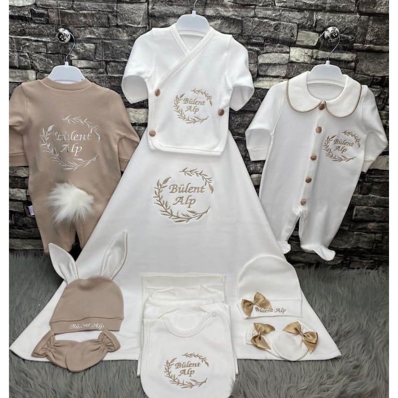 May include: A selection of baby clothes and accessories in cream and beige. The set includes a romper with a fluffy tail, a wrap top, a blanket, a bib, a hat, and mittens. Each item is embroidered with the text "B&uuml;lent Alp" within a wreath.