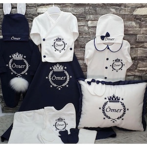 Peut inclure: Ensemble de vêtements et accessoires pour bébé, bleu marine et blanc. L'ensemble comprend un chapeau oreilles de lapin, des barboteuses, un bavoir, un oreiller et une couverture, tous avec le nom "Ömer" et un motif de couronne. Présenté sur fond neutre.