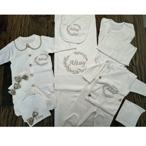 May include: A set of white baby clothes, featuring a onesie, bib, hat, and mittens, all with gold detailing and the name "Altay" embroidered. The collection also includes a blanket and a long-sleeved top.