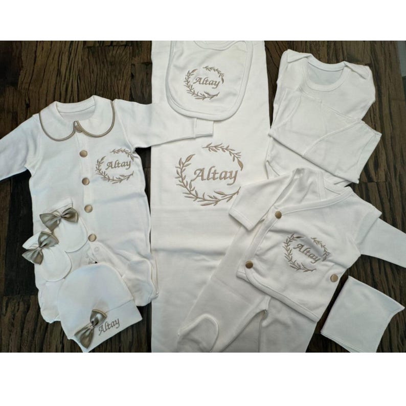 May include: A set of white baby clothes, featuring a onesie, bib, hat, and mittens, all with gold detailing and the name "Altay" embroidered. The collection also includes a blanket and a long-sleeved top.