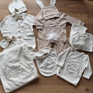 May include: A selection of baby clothes and accessories in cream and beige. The set includes a bunny-eared onesie, a blanket, a bib, a hat, a bow tie, mittens, and a wrap-over shirt. Each piece is embroidered with the name "Abdullah" within a wreath.