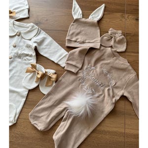 May include: A set of baby clothes in cream and tan. The set includes a cream onesie, a tan bunny hat, mittens, and a tan onesie with a white feather tail. Each item is embroidered with the name "Ismail".