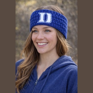 May include: A blue knit headband with the white letter "D" from Duke University. The headband is worn by a person wearing a blue hooded sweatshirt. The background is blurred with natural tones.