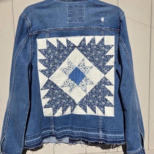 Puede incluir: Chaqueta vaquera azul con un cuadrado de patchwork decorativo en la espalda. El cuadrado de patchwork presenta un fondo blanco con motivos florales y geométricos azules. La chaqueta tiene un aspecto desgastado con bordes deshilachados y un pequeño rasgón cerca del hombro.