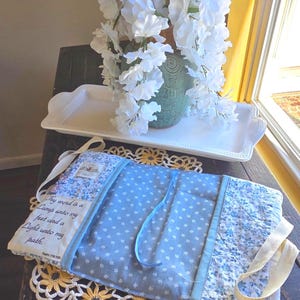 May include: A quilted fabric Bible cover in light blue with a white floral pattern, featuring a white fabric panel printed with the text: Thy word is a Lamp unto my feet and a Light unto my path. Psalm 119:105. It includes white fabric handles.