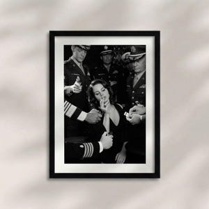 Puede incluir: Impresión enmarcada en blanco y negro que presenta a una mujer con un vestido negro fumando un cigarrillo, rodeada de hombres con uniformes militares. La imagen tiene una estética vintage de cine negro.