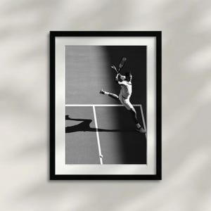 May include: Black and white framed print of a tennis player mid-swing. The player is wearing a white uniform and is positioned on a tennis court. The image captures the dynamic action of the sport, with the player's shadow visible on the court.