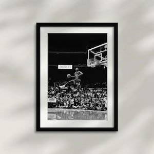 以下が含まれることがあります： バスケットボール選手が空中を飛び、ボールをダンクしようとしている白黒の額入りプリント。画像は観客で賑わうスタジアムで撮影されています。「BUSCH」の文字が看板に見えます。