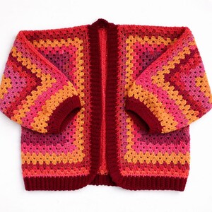 May include: A handmade, open-front crochet cardigan in a vibrant color palette. The sweater features a granny square design with shades of red, pink, and orange. The sleeves and trim are a deep burgundy. This cozy garment is perfect for layering.