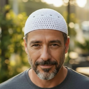 Può includere: Un berretto bianco all'uncinetto con un design traforato. Il berretto è indossato da una persona con barba grigia e una maglietta grigia. Lo sfondo è sfocato con la luce naturale.