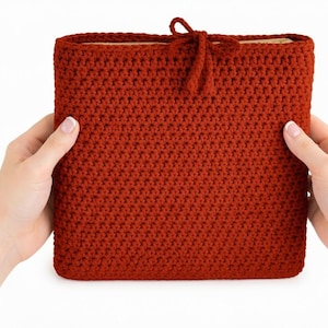 Pode incluir: Uma capa de livro de crochê em um tom quente de marrom avermelhado. A capa é retangular com um padrão de ponto texturizado e um pequeno fecho de laço na parte superior. O item feito à mão é segurado por duas mãos.