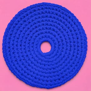 May include: A round, blue crocheted item with a central hole. The item is made of thick yarn and has a textured surface. The background is a solid pink color. This could be a trivet or a coaster.