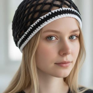 May include: Black crochet beanie with a white and black striped brim. The hat features a net-like design and is worn on a person with blonde hair and blue eyes. The beanie is a stylish accessory.