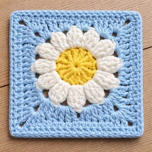 May include: A square, crocheted daisy motif. The center of the flower is yellow, surrounded by white petals. The background is a light blue, square shape with a textured stitch pattern. The daisy is centered within the blue square.