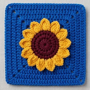 May include: A square, crocheted piece featuring a sunflower design. The square is a vibrant blue, with a yellow sunflower and a deep burgundy center. The crochet stitches are clearly visible, adding texture to the handmade item.