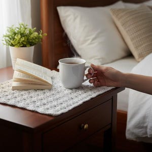 May include: A white ceramic mug filled with a steaming hot beverage sits next to an open book on a light gray, textured, rectangular crochet mat. The mat is placed on a dark wooden surface, providing a neutral base for the items displayed.