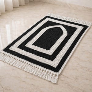 May include: A rectangular black and white crochet prayer rug with a central arched design. The rug features a black field framed by white borders and a fringe along the short edges. The rug is placed on a marble floor.