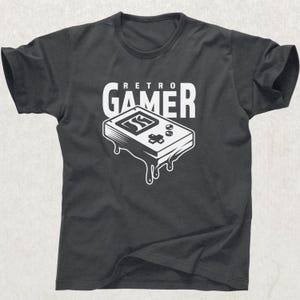 May include: Dark gray t-shirt featuring a white graphic of a retro handheld gaming device with the text "RETRO GAMER" above it. The game console graphic has a melting effect, giving it a unique look. A classic design for gamers.
