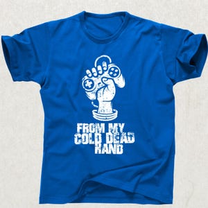Pode incluir: Camiseta azul royal com um gráfico branco de uma mão segurando um controle de videogame. O texto "FROM MY COLD DEAD HAND" é impresso abaixo em uma fonte desgastada. Uma camiseta casual para jogadores.
