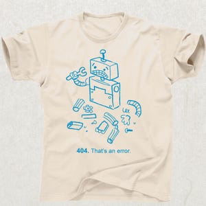 May include: Cream-colored t-shirt featuring a distressed blue illustration of a robot holding a wrench, surrounded by scattered parts. The text "404. That's an error." is printed below the robot. The shirt has a crew neck and short sleeves.