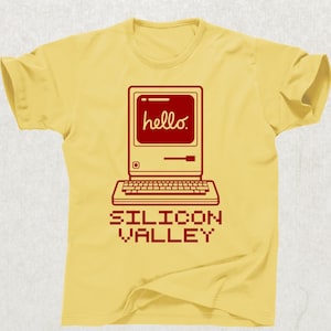 May include: A yellow t-shirt featuring a red retro computer graphic with the word "hello" on the screen. Below the computer is the text "SILICON VALLEY" in a pixelated font. The shirt has short sleeves and a crew neck.