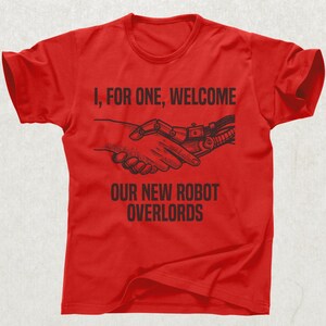 May include: Red t-shirt with black text and graphic. The text reads "I, FOR ONE, WELCOME OUR NEW ROBOT OVERLORDS" above a graphic of a human hand shaking a robotic hand. The shirt has short sleeves and a crew neck.