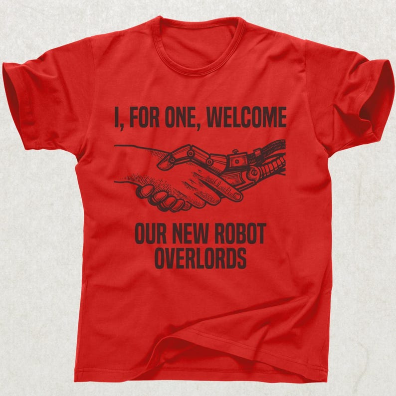 May include: Red t-shirt with black text and graphic. The text reads "I, FOR ONE, WELCOME OUR NEW ROBOT OVERLORDS" above a graphic of a human hand shaking a robotic hand. The shirt has short sleeves and a crew neck.