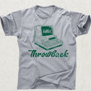 Könnte beinhalten: Graues T-Shirt mit einem grünen Aufdruck eines Retro-Computers mit dem Wort "hello" auf dem Bildschirm. Darunter steht das Wort "Throwback" in einer Schreibschrift. Das T-Shirt hat kurze Ärmel und einen Rundhalsausschnitt.