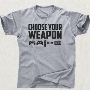Pode incluir: Camiseta cinza mesclada com a frase "CHOOSE YOUR WEAPON" em letras pretas em negrito. Abaixo, ilustrações pretas de um controle de jogo, um gamepad, um controle remoto, um console de jogos e um teclado.