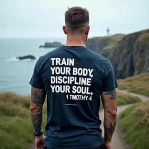 Puede incluir: Una camiseta azul marino con texto blanco: "TRAIN YOUR BODY, DISCIPLINE YOUR SOUL, 1 TIMOTHY 4." La persona que lleva la camiseta está de pie cerca del mar, con un camino y un faro al fondo.