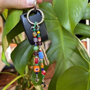 Puede incluir: Un llavero de coche negro con una llave plateada y un llavero de cuentas. El llavero presenta cuentas de vidrio de colores en varias formas y tamaños, incluyendo redondas, cuadradas y oblongas. Las cuentas son en tonos de rojo, azul, verde, amarillo y morado.