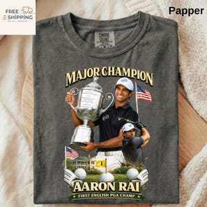 May include: Dark grey t-shirt with a graphic of golfer Aaron Rai holding a trophy, text reading "MAJOR CHAMPION" and "AARON RAI FIRST ENGLISH PGA CHAMP". Features American flags and golf balls.