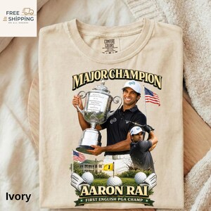 May include: Ivory t-shirt with a golf-themed graphic. The design includes a golfer holding a trophy, the text "MAJOR CHAMPION" and "AARON RAI FIRST ENGLISH PGA CHAMP." The image also features golf balls and the American flag.