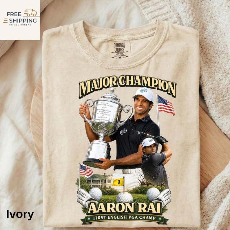 May include: Ivory t-shirt with a golf-themed graphic. The design includes a golfer holding a trophy, the text "MAJOR CHAMPION" and "AARON RAI FIRST ENGLISH PGA CHAMP." The image also features golf balls and the American flag.