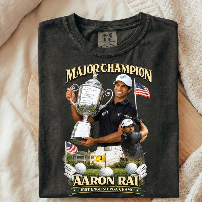 May include: Black Comfort Colors t-shirt with a graphic of golfer Aaron Rai holding a trophy, text reading "MAJOR CHAMPION" and "AARON RAI FIRST ENGLISH PGA CHAMP". Includes golf balls, gloves, and an American flag.