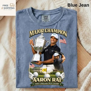 May include: Blue jean coloured t-shirt with a graphic of golfer Aaron Rai holding a trophy. The text "MAJOR CHAMPION" is above the image, and "AARON RAI FIRST ENGLISH PGA CHAMP" is below. The shirt also has the Comfort Colors brand label.