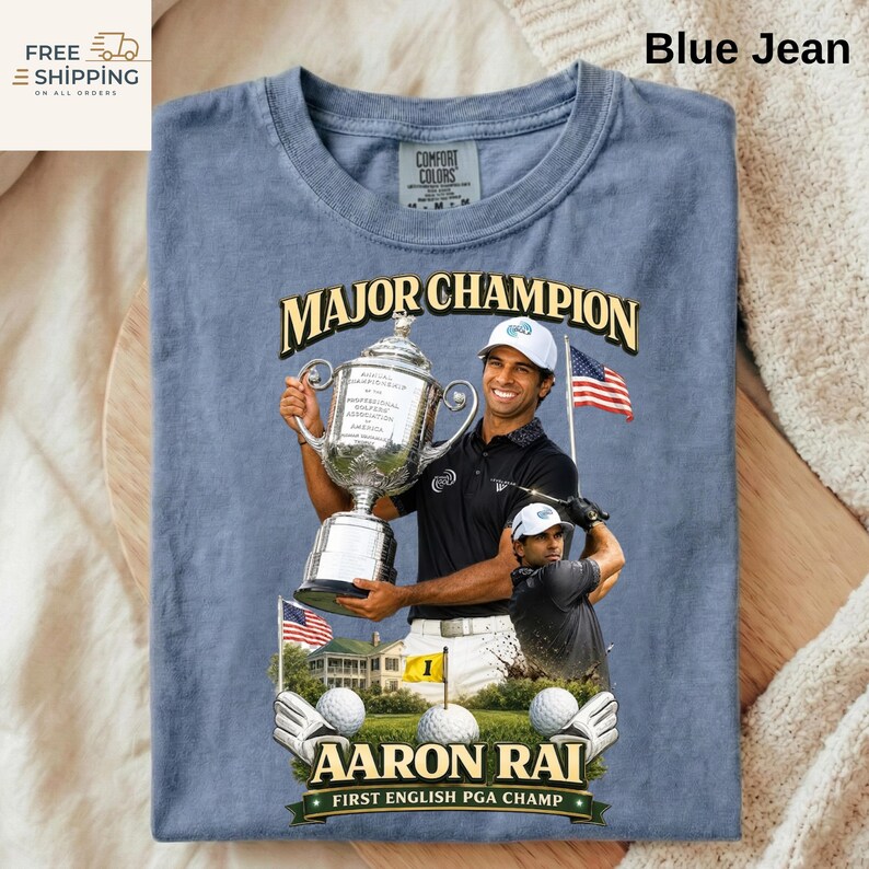 May include: Blue jean coloured t-shirt with a graphic of golfer Aaron Rai holding a trophy. The text "MAJOR CHAMPION" is above the image, and "AARON RAI FIRST ENGLISH PGA CHAMP" is below. The shirt also has the Comfort Colors brand label.