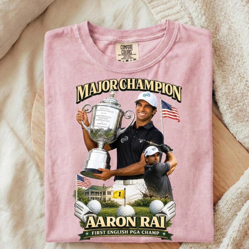 May include: Pink Comfort Colors t-shirt with a graphic of a golfer holding a trophy, the text "MAJOR CHAMPION" and "AARON RAI FIRST ENGLISH PGA CHAMP". Features golf balls, flags, and a golf course scene.